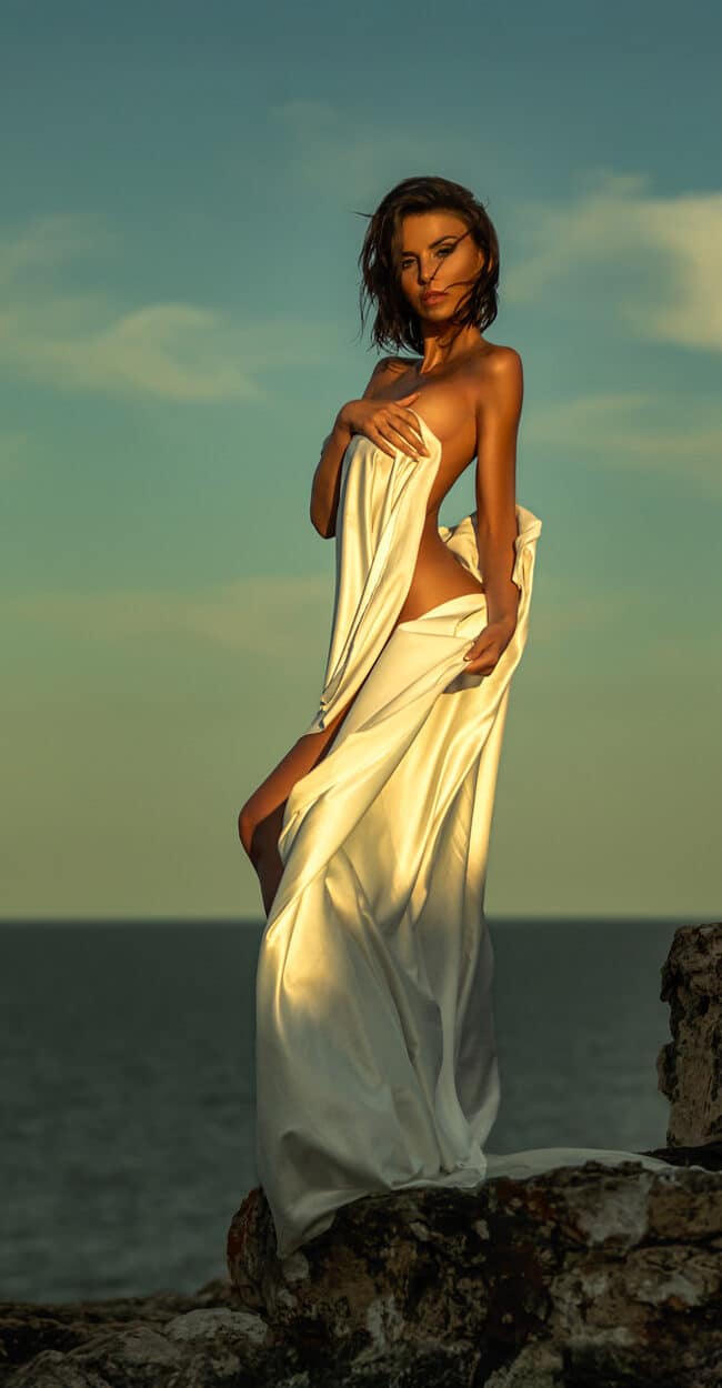Fine-art portrait by the sea in Portixol, Palma de Mallorca – model wrapped in cream silk on the rocks at sunset