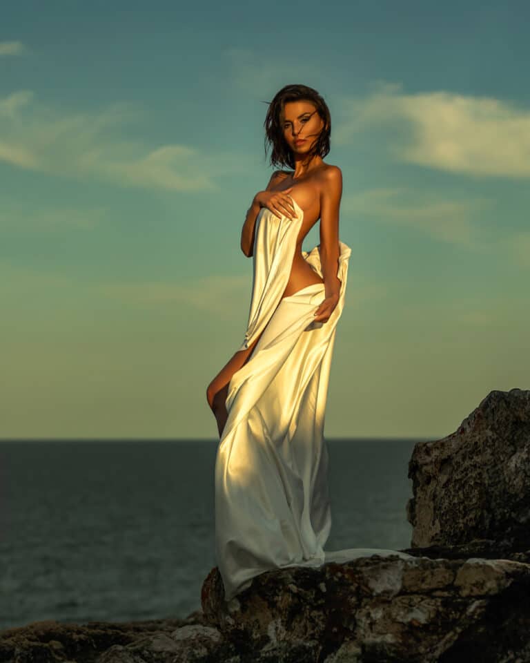 Fine-art portrait by the sea in Portixol, Palma de Mallorca – model wrapped in cream silk on the rocks at sunset
